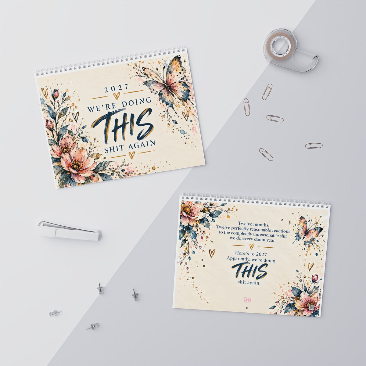 Two spiral-bound desk calendars with floral and butterfly designs are arranged on a light gray surface with tape, paper clips, and pins around them. The calendars feature the text '2027 WE'RE DOING THIS SHIT AGAIN' and 'Twelve months.