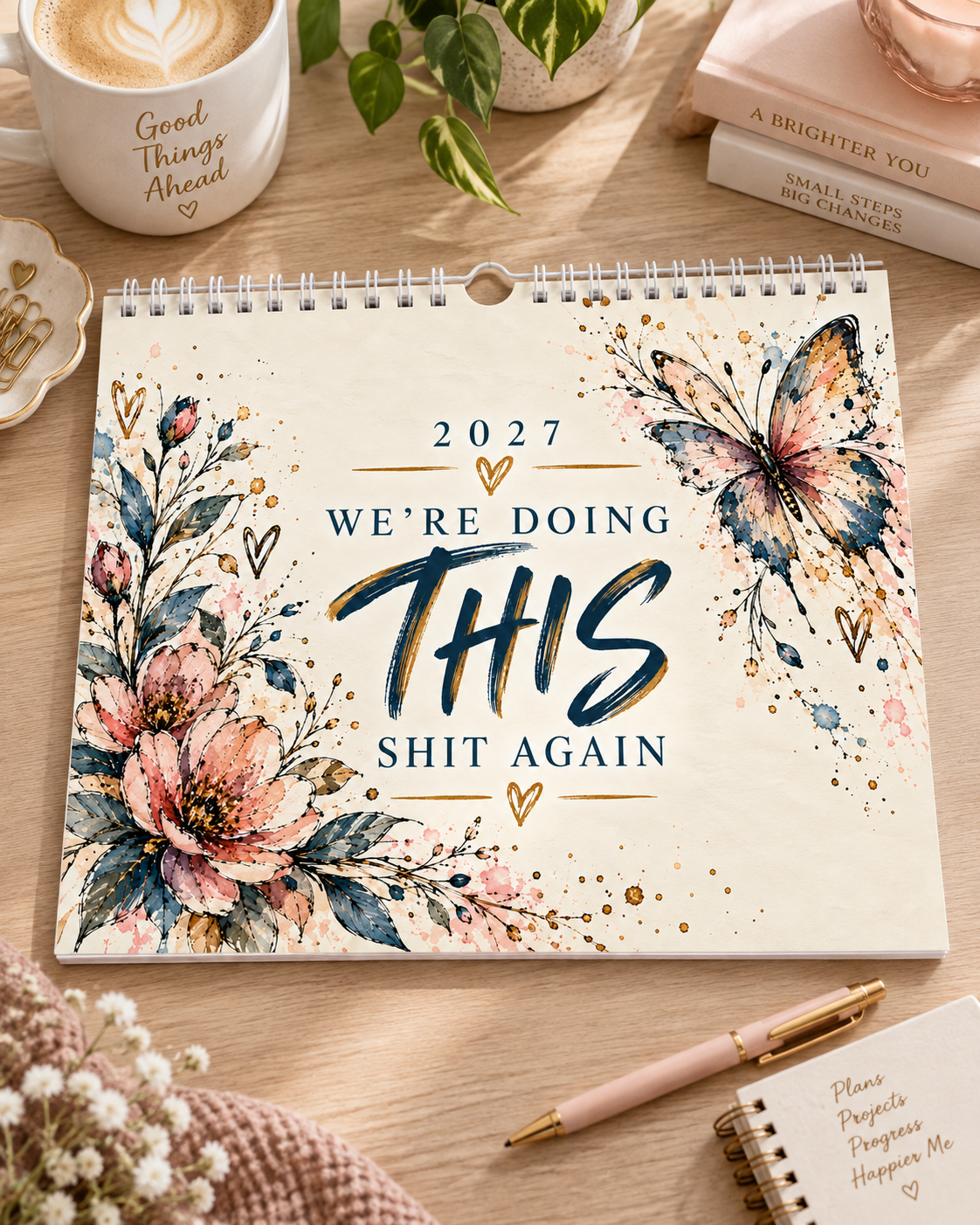 A spiral-bound 2027 desk calendar with a humorous quote reading 'WE'RE DOING THIS SHIT AGAIN' in navy lettering, surrounded by watercolor floral illustrations and a butterfly. The calendar is styled on a wooden desk with a coffee mug, books, a pen, and a small notebook.
